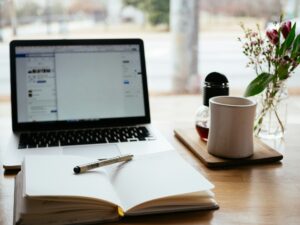 Laptop in the Background for web development along with a mug of coffee by its side and a notebook with a pen on it.