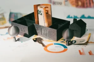 A conceptual image representing content marketing ROI, featuring miniature house models, a euro currency note rolled inside a model building, a metal key, and analytical charts with pie and bar graphs in the background, symbolizing investment returns and financial analysis.