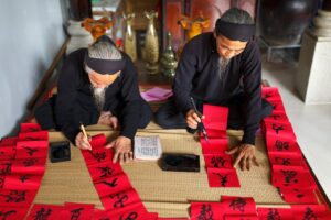 Two elderly men practicing traditional calligraphy on red paper, illustrating the deep connection between language and culture through handwritten scripts and cultural rituals.