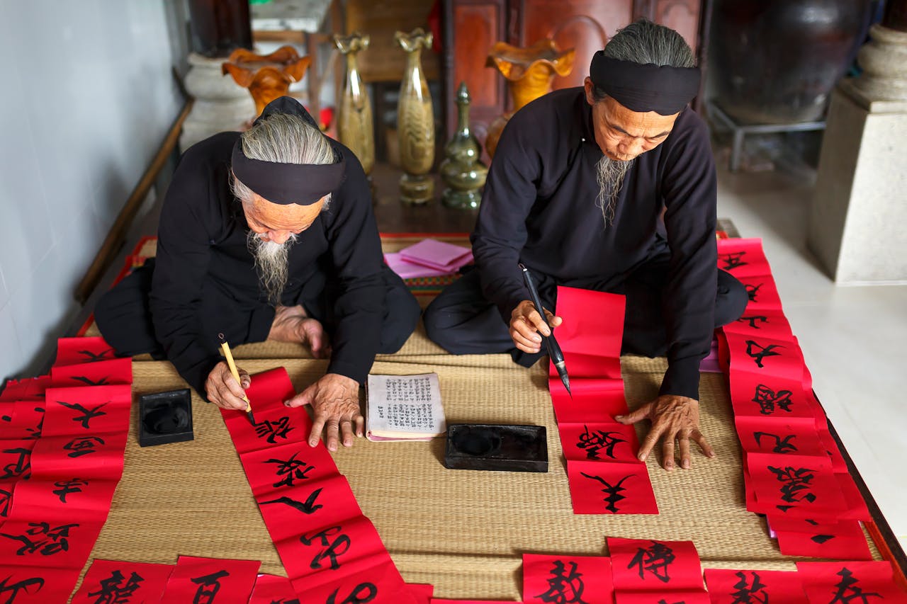 Two elderly men practicing traditional calligraphy on red paper, illustrating the deep connection between language and culture through handwritten scripts and cultural rituals.