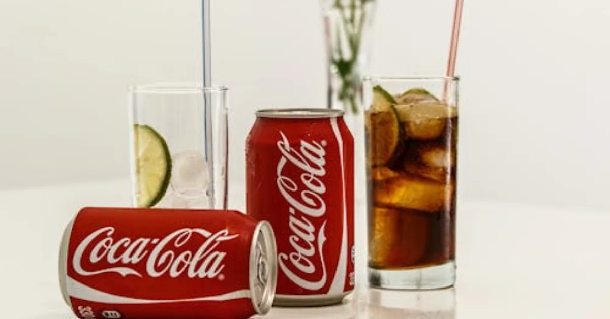 “Three Coca-Cola cans and glasses with straws on a table, including one filled with ice and a lemon slice
